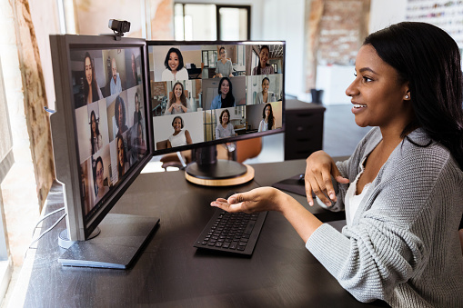 Das Bild zeigt eine junge Frau, die mit ihren Kolleg:innen eine Videokonferenz online an ihrem Computer durchführt. Sie sitzt vor Ihren Bildschirmen, um das pandemiebedingte Social Distancing auch auf Berufswegen einzuhalten.